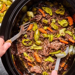 Two hands use forks to shred cooked pot roast with sliced carrots, celery, and pepperoncini peppers in a slow cooker. Bowls of pickled vegetables are visible nearby.