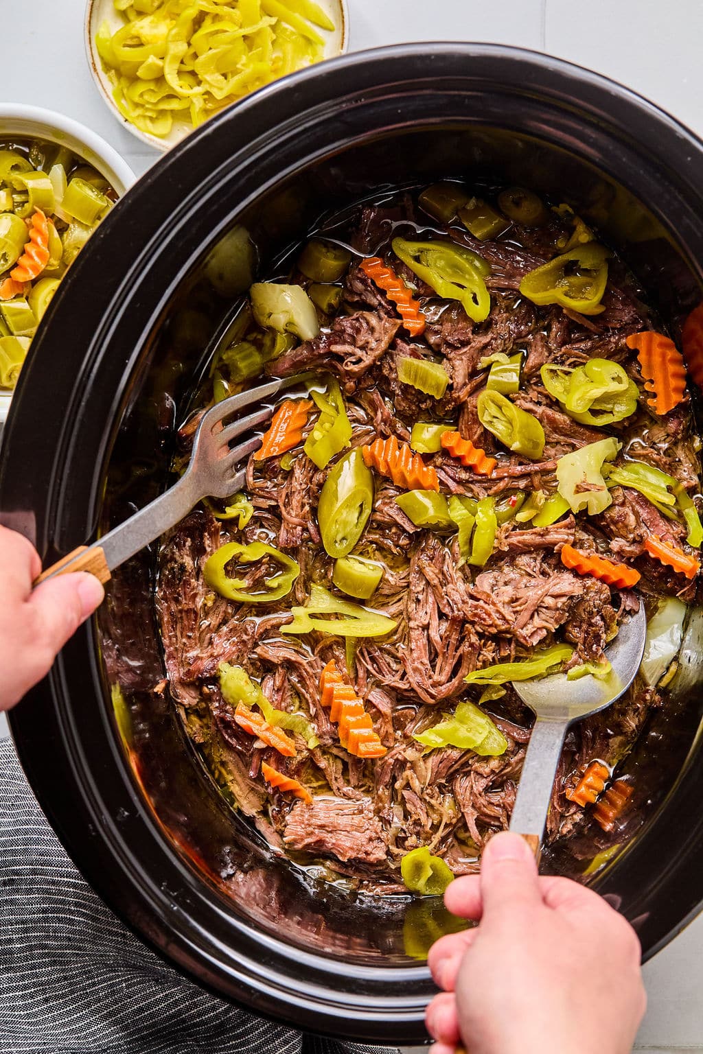 Two hands use forks to shred cooked pot roast with sliced carrots, celery, and pepperoncini peppers in a slow cooker. Bowls of pickled vegetables are visible nearby.