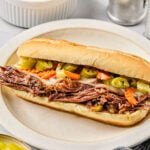 A hoagie roll filled with shredded meat and topped with pickled vegetables sits on a plate, with small bowls of pickled vegetables nearby.
