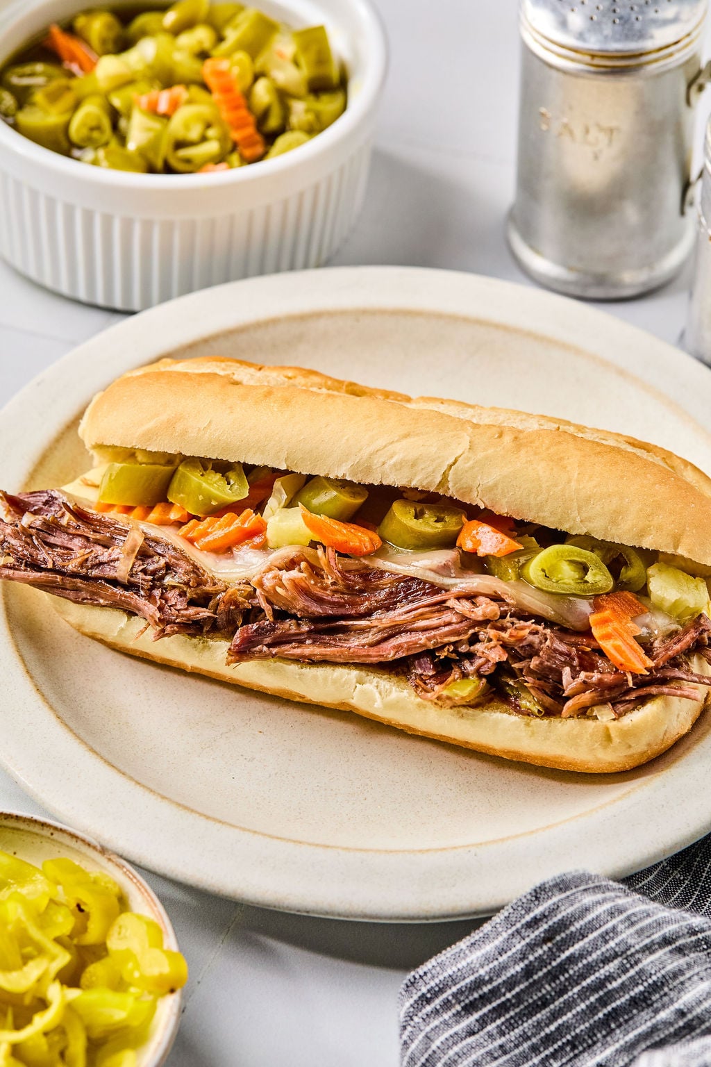 A hoagie roll filled with shredded meat and topped with pickled vegetables sits on a plate, with small bowls of pickled vegetables nearby.