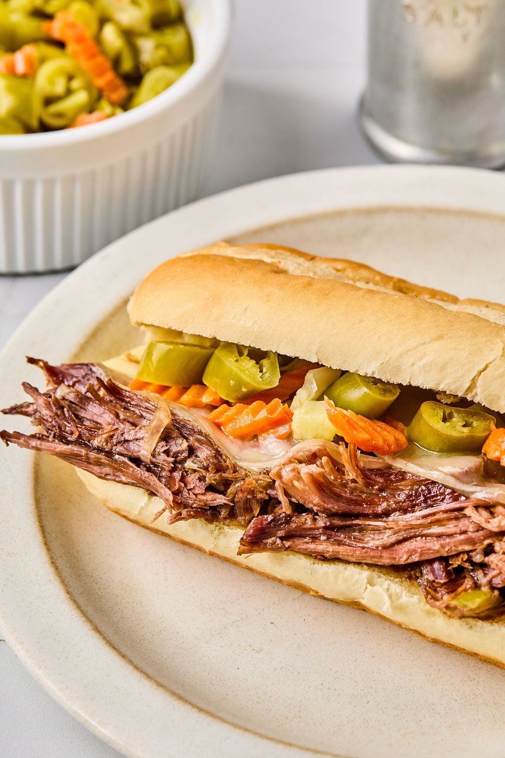 A sandwich with shredded beef, sliced pickled peppers, and carrots on a hoagie roll sits on a plate. A dish of pickled vegetables is in the background.