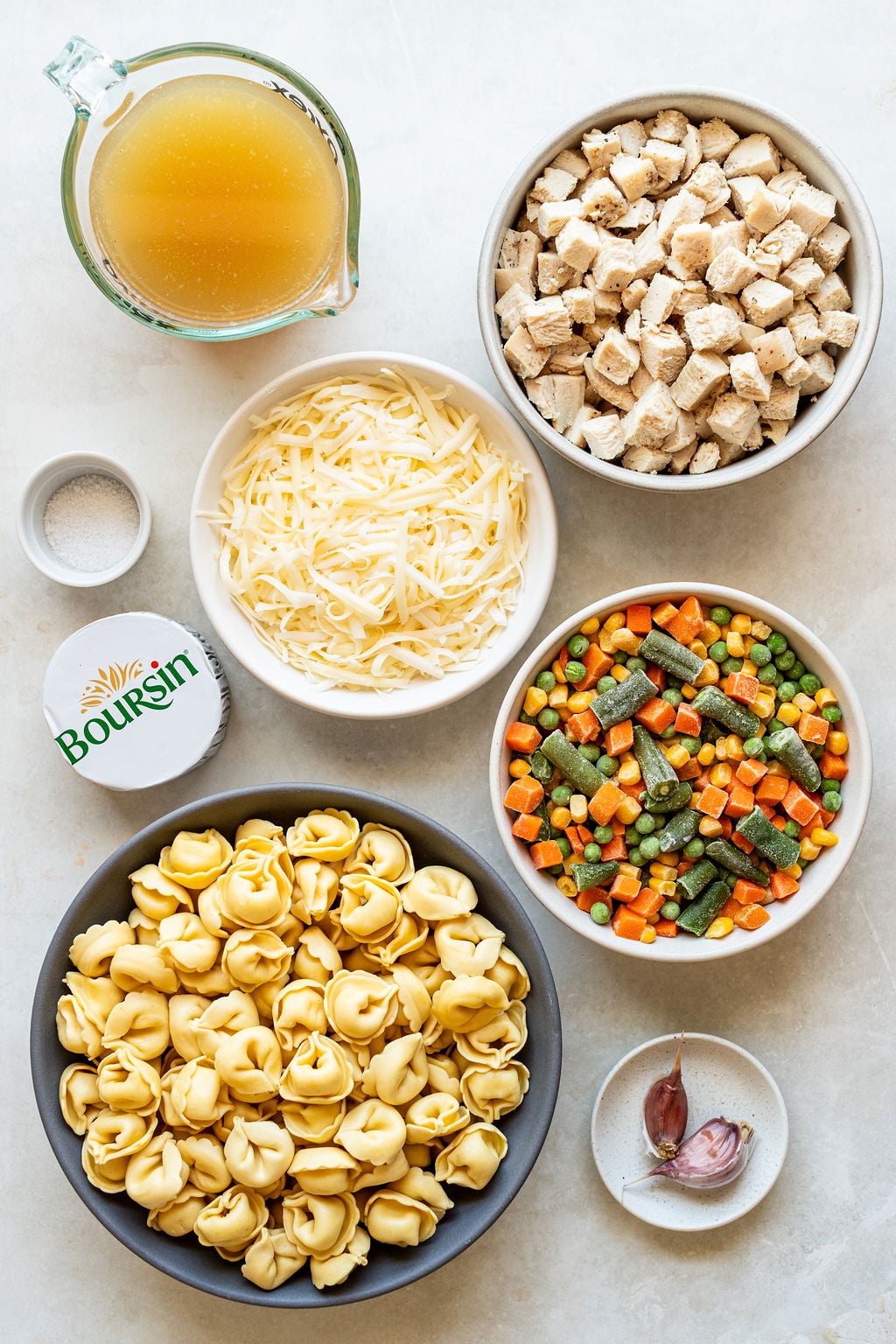 Top view of ingredients including cubed chicken, shredded cheese, mixed vegetables, tortellini, garlic cloves, Boursin cheese, chicken broth, and a small bowl of salt on a white surface.