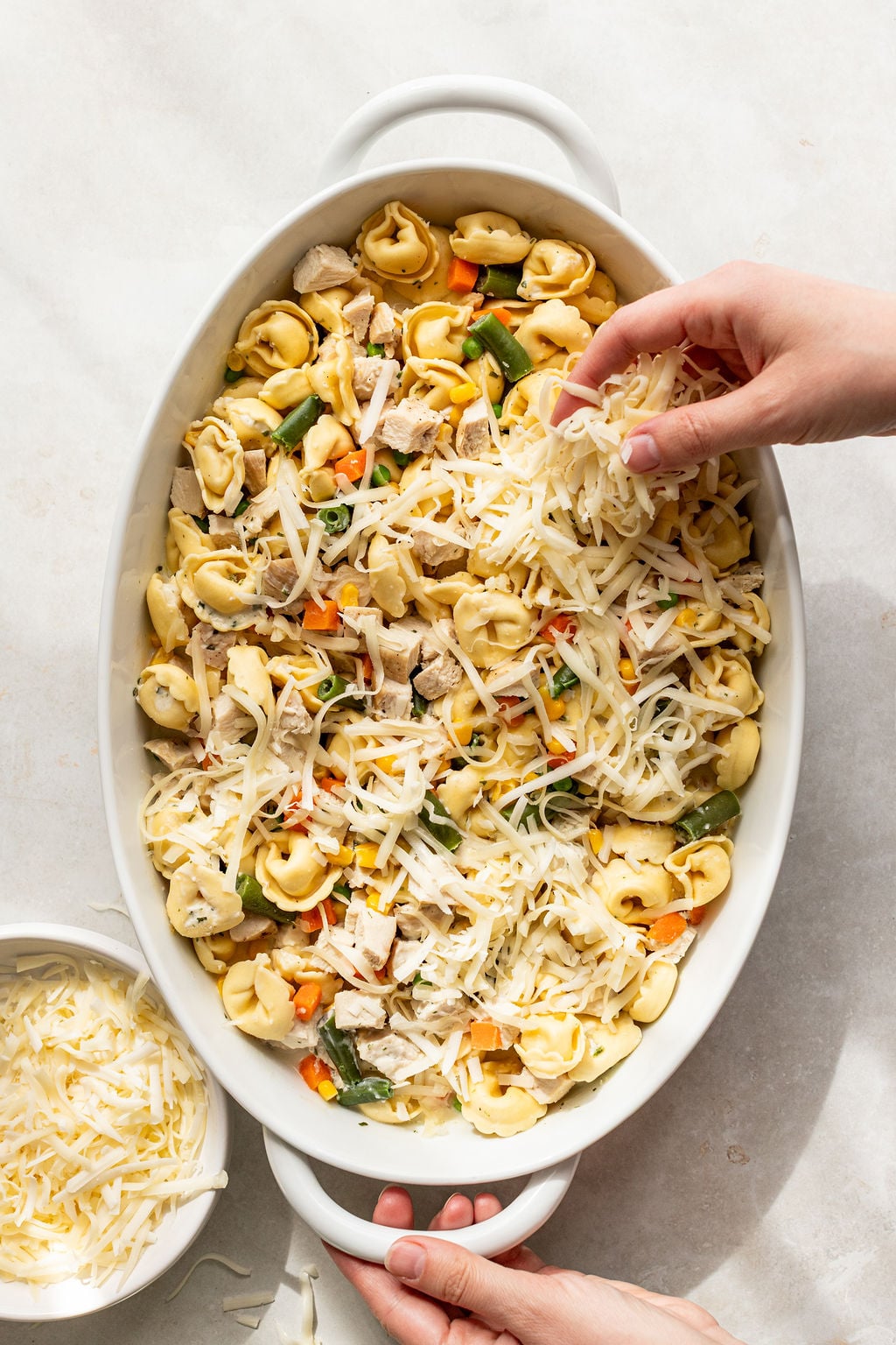 A persons hands sprinkle shredded cheese over a casserole dish filled with tortellini, vegetables, and pieces of chicken.