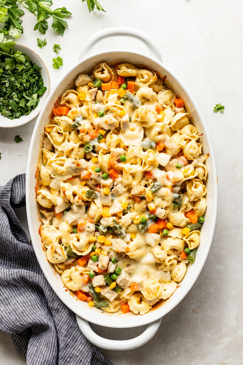 Baked tortellini casserole with mixed vegetables, melted cheese on top, in a white oval dish, next to a bowl of chopped parsley and a striped cloth napkin.