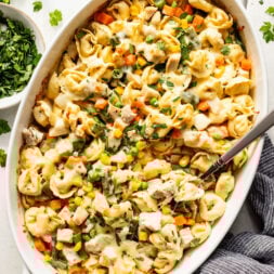 Baked tortellini casserole with mixed vegetables, chicken pieces, and melted cheese in a white oval dish with a serving spoon and fresh herbs on the side.