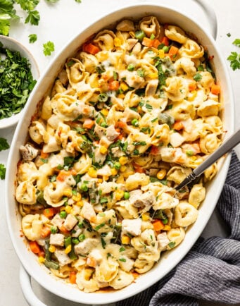 Baked tortellini casserole with mixed vegetables, chicken pieces, and melted cheese in a white oval dish with a serving spoon and fresh herbs on the side.
