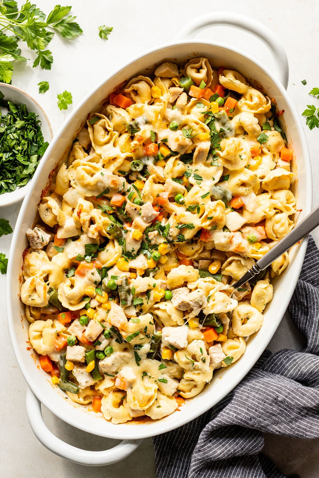 Baked tortellini casserole with mixed vegetables, chicken pieces, and melted cheese in a white oval dish with a serving spoon and fresh herbs on the side.