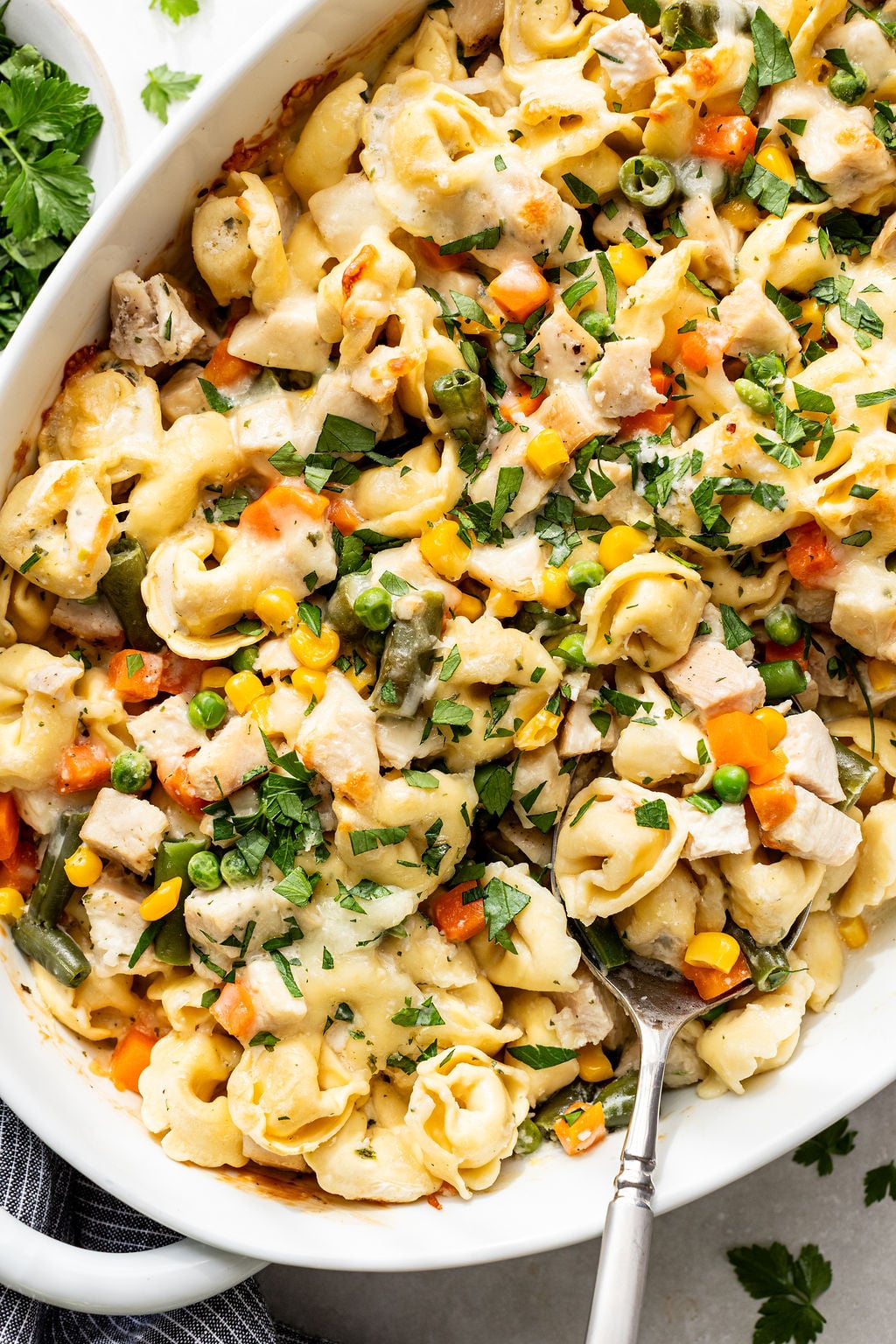 Baked tortellini casserole with mixed vegetables, chicken, melted cheese, and fresh herbs in a white dish with a serving spoon.