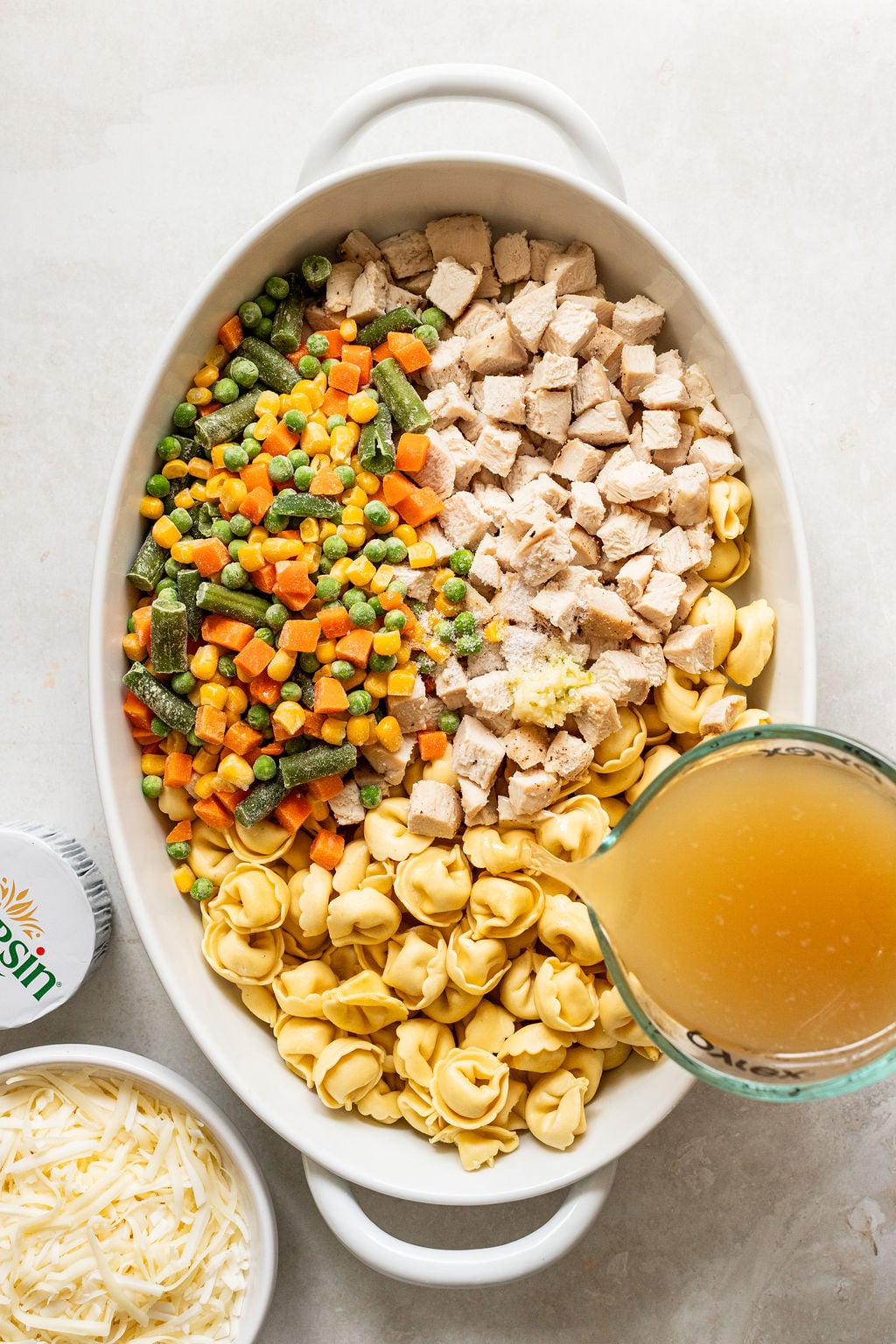 Oval baking dish with uncooked tortellini, diced cooked chicken, mixed frozen vegetables, shredded cheese, and a measuring cup pouring broth.