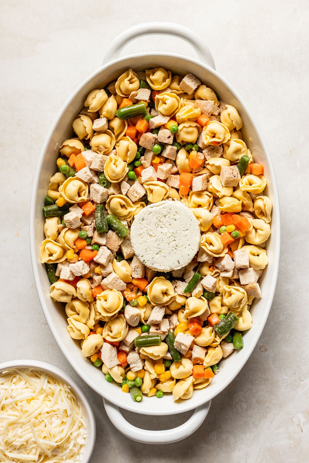 A white oval baking dish filled with tortellini, mixed vegetables, cubed chicken, and a round block of seasoned cream cheese in the center. A bowl of shredded cheese is beside the dish.