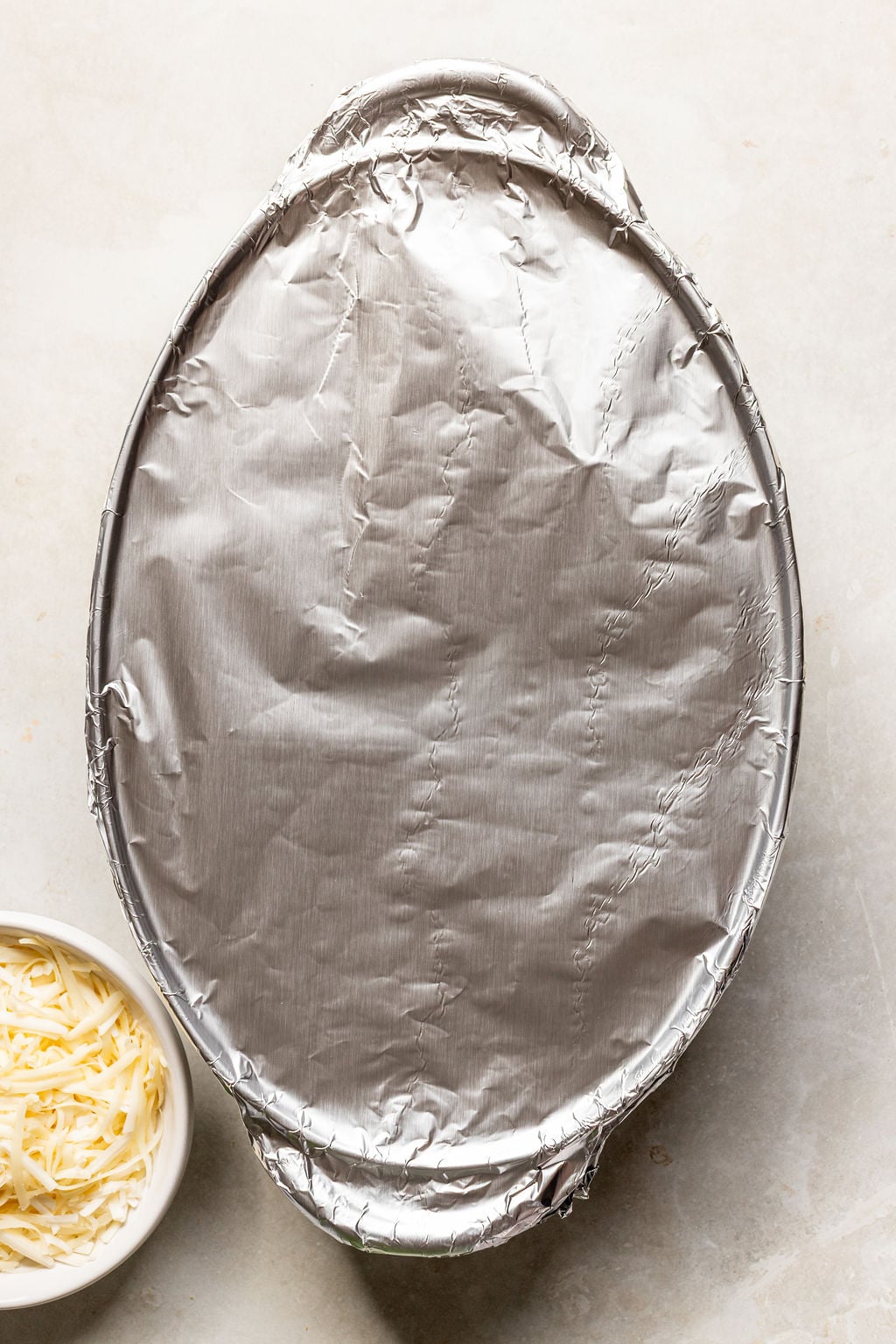 An oval baking dish covered with aluminum foil sits on a light surface next to a small bowl of shredded cheese.
