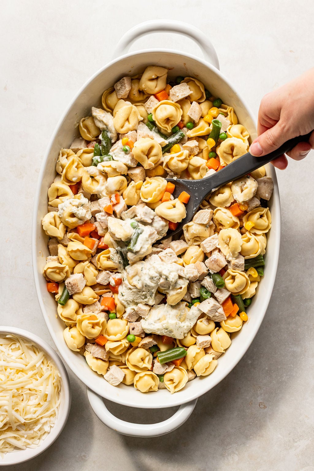 An oval baking dish filled with tortellini, diced chicken, mixed vegetables, and creamy sauce being stirred with a black spoon; a bowl of shredded cheese sits nearby.