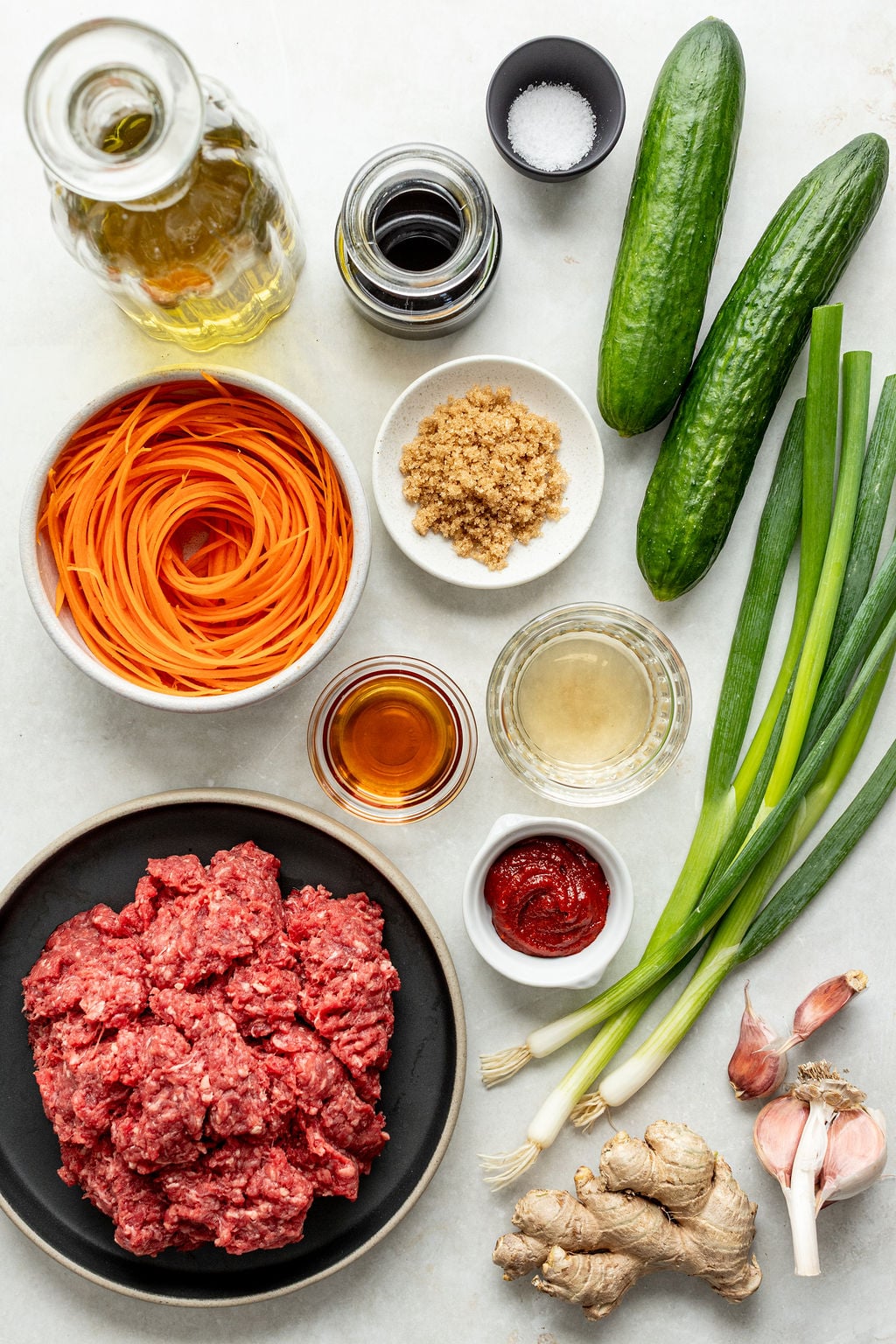 A flat lay of ingredients including ground beef, cucumbers, green onions, shredded carrots, ginger, garlic, shallot, brown sugar, tomato paste, sauces, salt, and oil on a white surface.