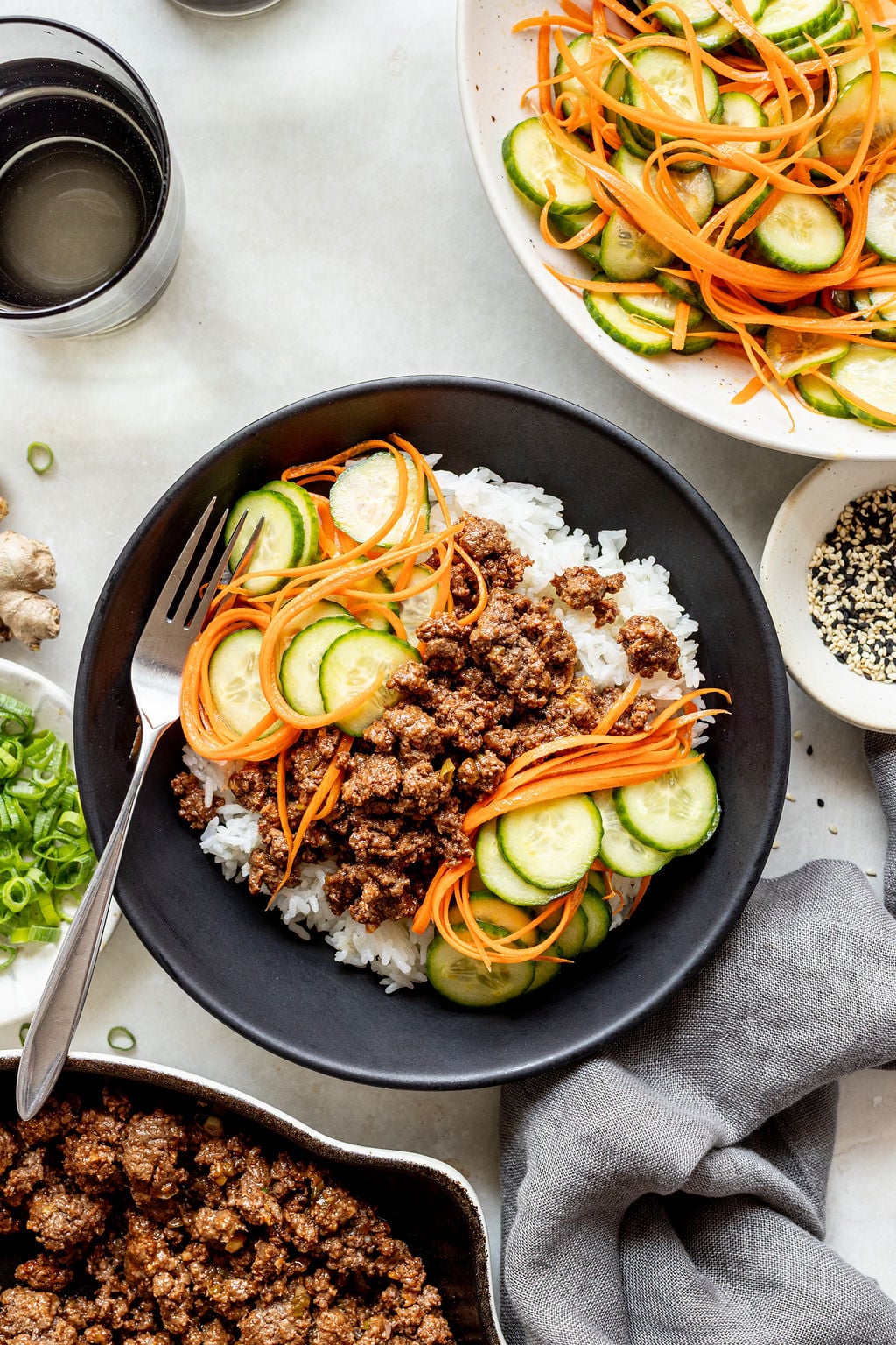 A black bowl filled with white rice, ground beef, sliced cucumbers, and julienned carrots, with a fork resting on the side. Other dishes and toppings are visible nearby.