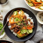 A bowl of white rice topped with ground meat, sliced cucumbers, shredded carrots, green onions, and sesame seeds, with a fork on the side and a salad in the background.