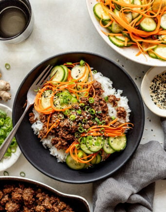 A bowl of white rice topped with ground meat, sliced cucumbers, shredded carrots, green onions, and sesame seeds, with a fork on the side and a salad in the background.