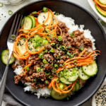 A bowl of white rice topped with ground meat, sliced cucumbers, shredded carrots, chopped green onions, and sesame seeds, with a fork on the side.