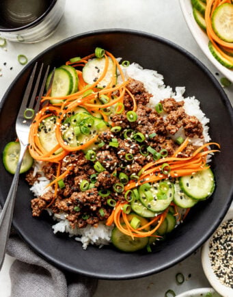 A bowl of white rice topped with ground meat, sliced cucumbers, shredded carrots, chopped green onions, and sesame seeds, with a fork on the side.