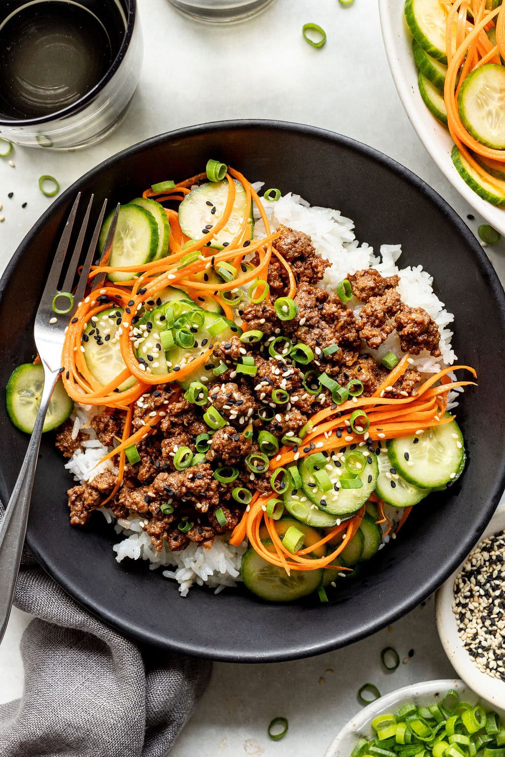 A bowl of white rice topped with ground meat, sliced cucumbers, shredded carrots, chopped green onions, and sesame seeds, with a fork on the side.