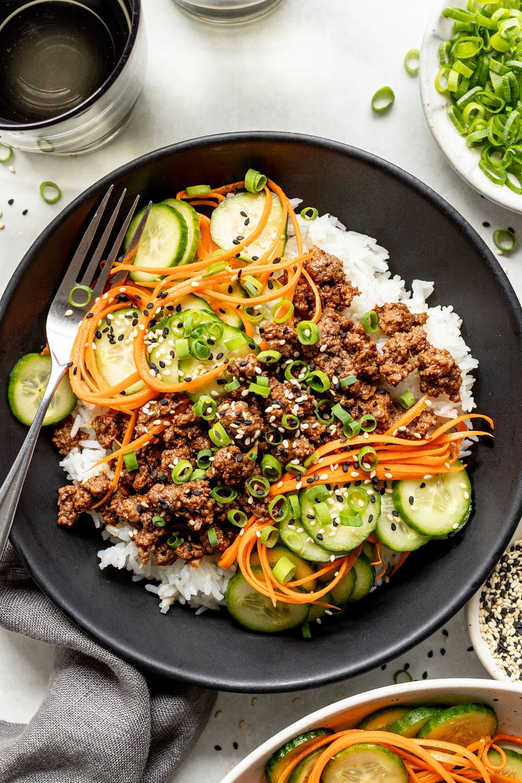 A black bowl filled with white rice, ground meat, sliced cucumbers, shredded carrots, chopped green onions, and black sesame seeds, with a fork on the side.
