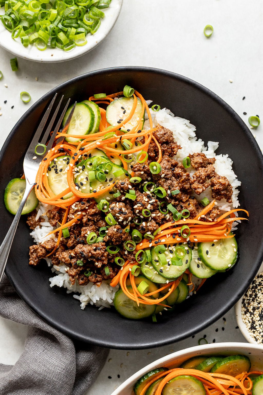 A black bowl filled with white rice, ground beef, sliced cucumbers, shredded carrots, chopped green onions, and black sesame seeds, with a fork resting inside.