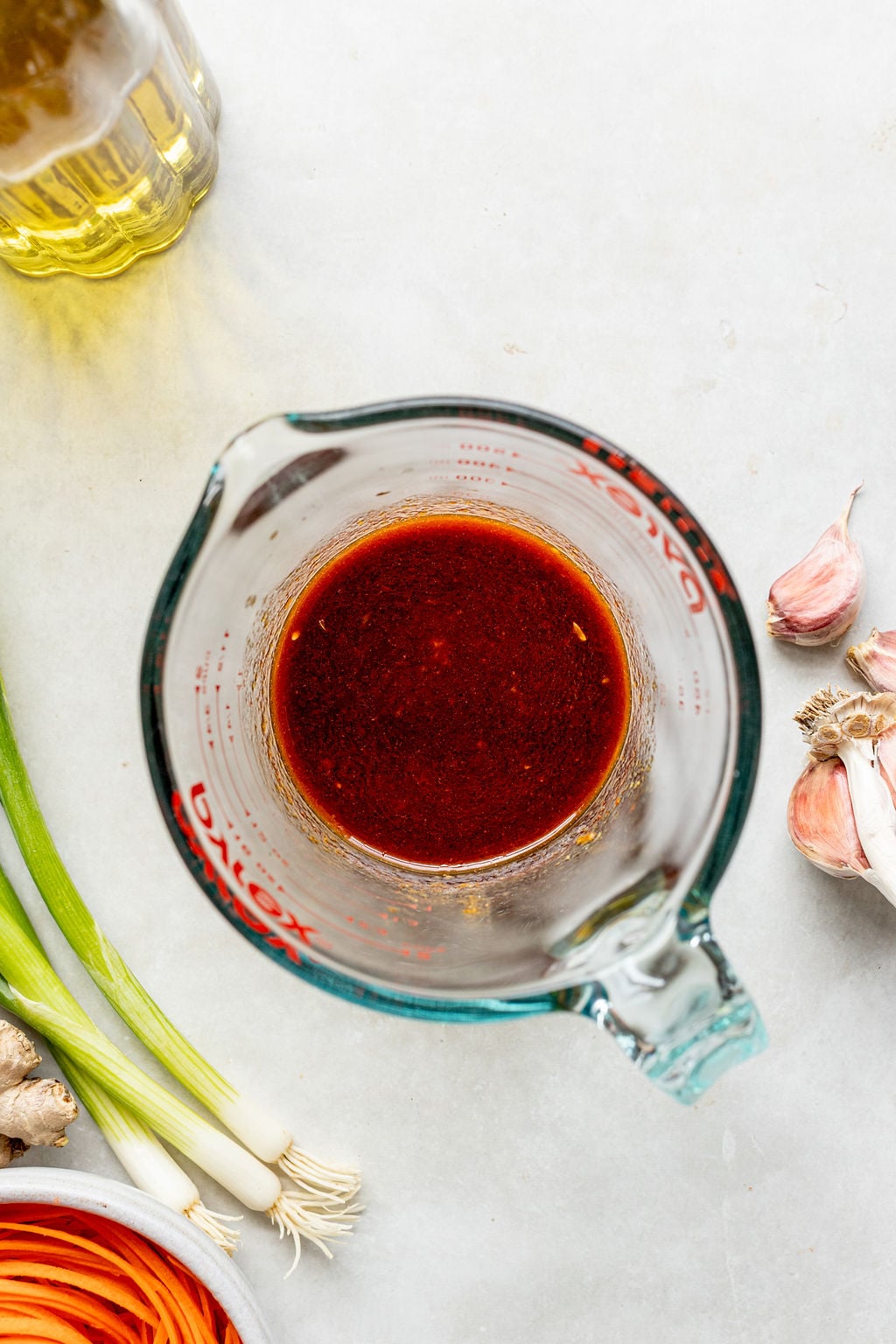 A glass measuring cup filled with a dark red liquid sits on a countertop surrounded by green onions, garlic, ginger, a bowl of sliced carrots, and a bottle of oil.