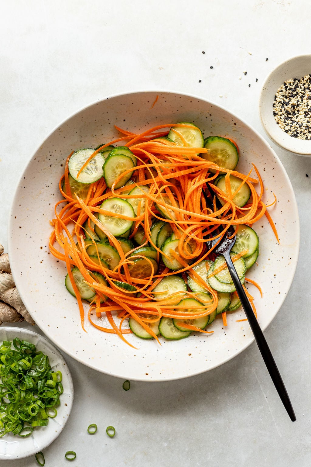 A bowl filled with sliced cucumbers and shredded carrots, with a black fork, sits next to small bowls of sliced green onions, grated ginger, and a seed mix.