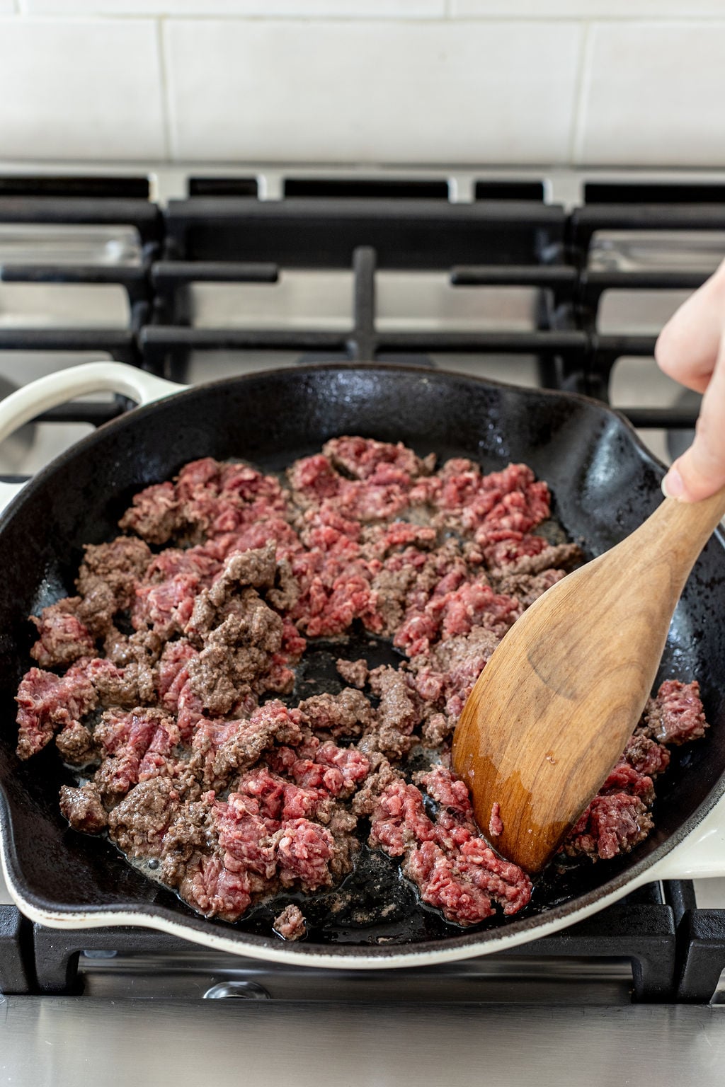 Ground beef being cooked in a cast iron skillet on a stovetop, with a wooden spatula stirring the meat.
