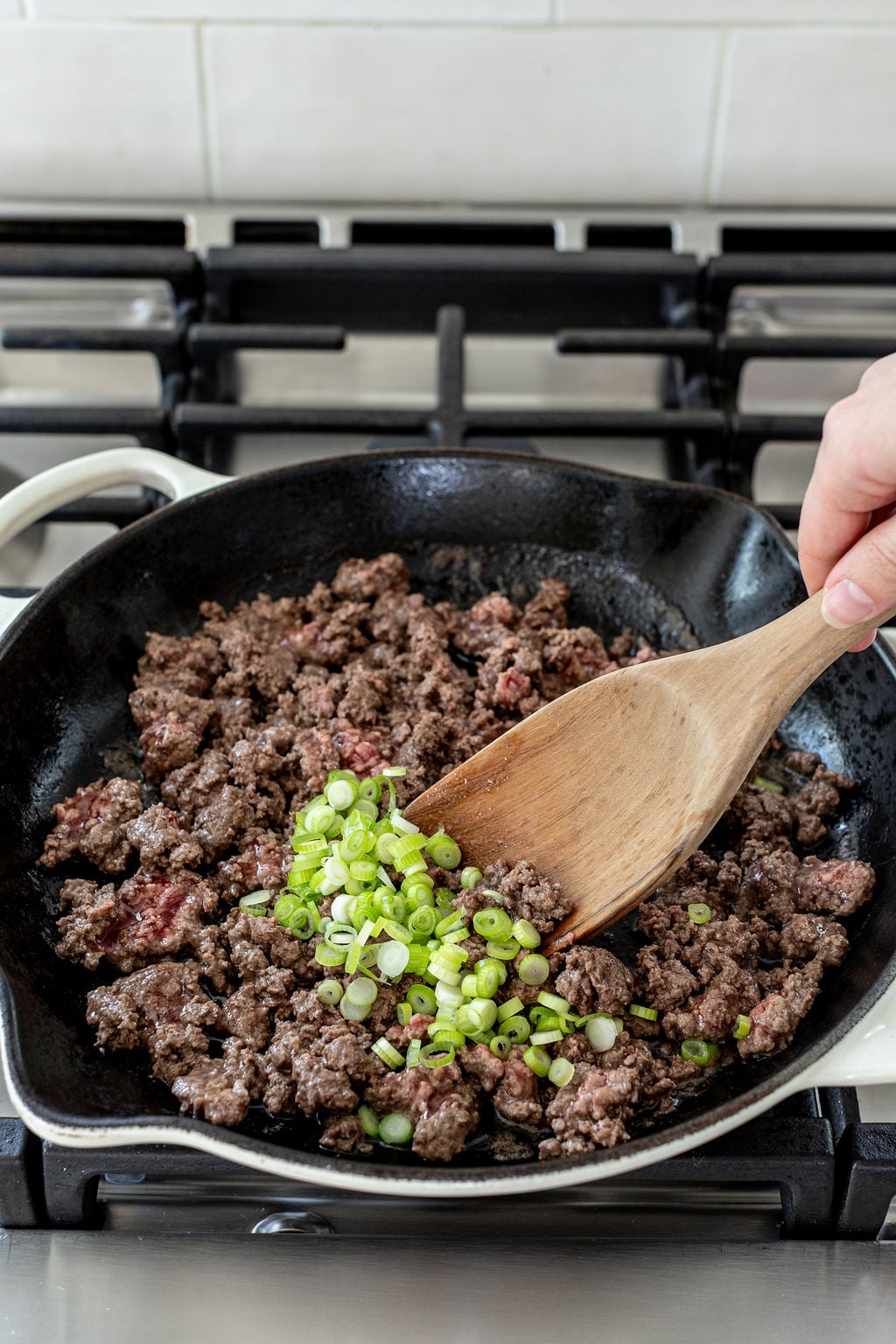 Ground beef is being cooked in a skillet on a stove, with chopped green onions being stirred in using a wooden spatula.