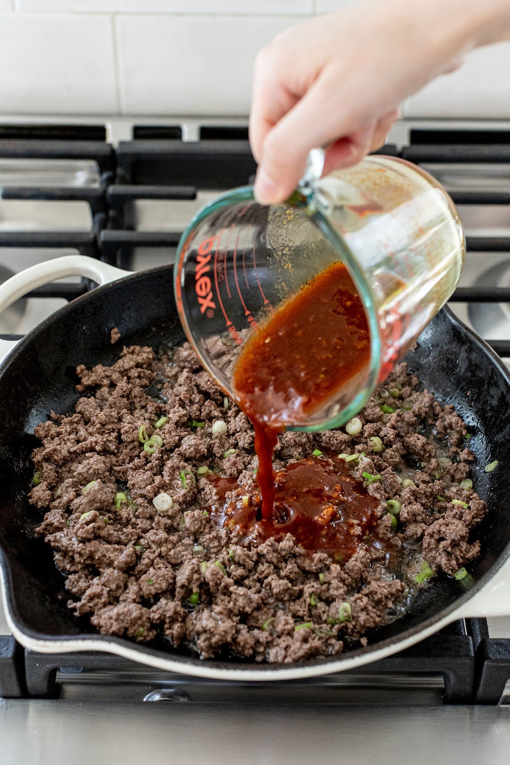 A hand pours red sauce from a measuring cup into a skillet with cooked ground beef and chopped green onions on a stovetop.