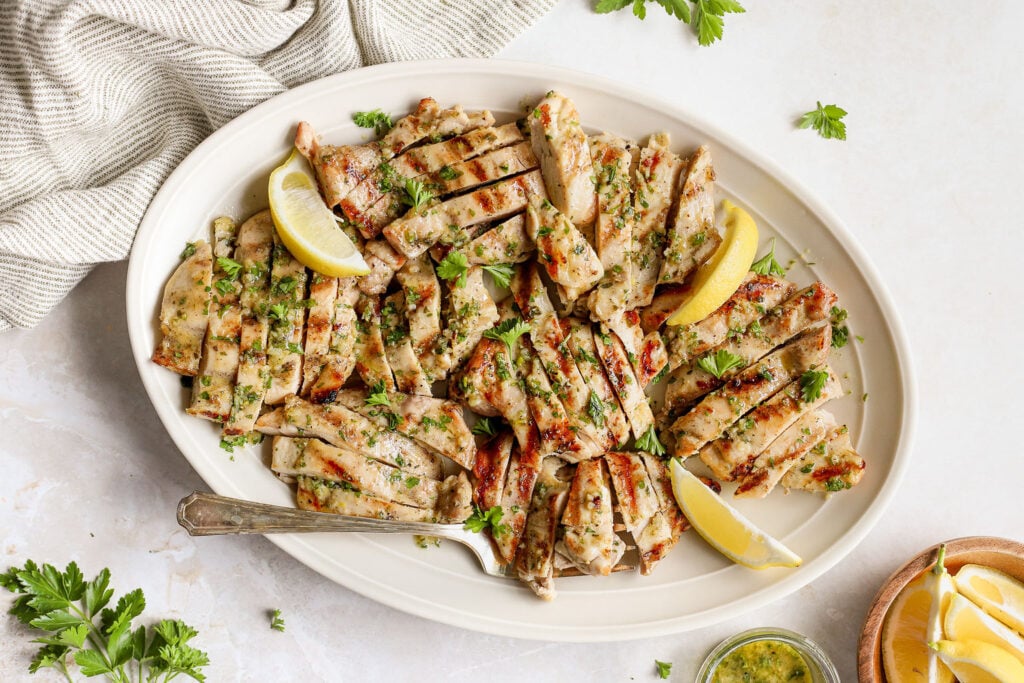 Sliced grilled chicken breast garnished with chopped herbs and lemon wedges, served on a white oval platter with a fork.