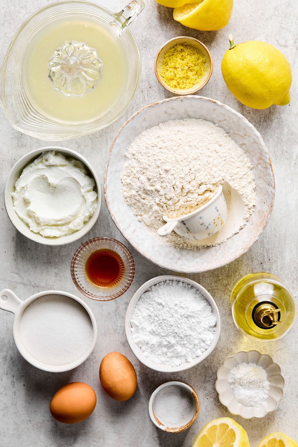 Assorted baking ingredients on a countertop, including flour, sugar, eggs, ricotta, oil, vanilla, lemons, lemon zest, baking powder, and powdered sugar.