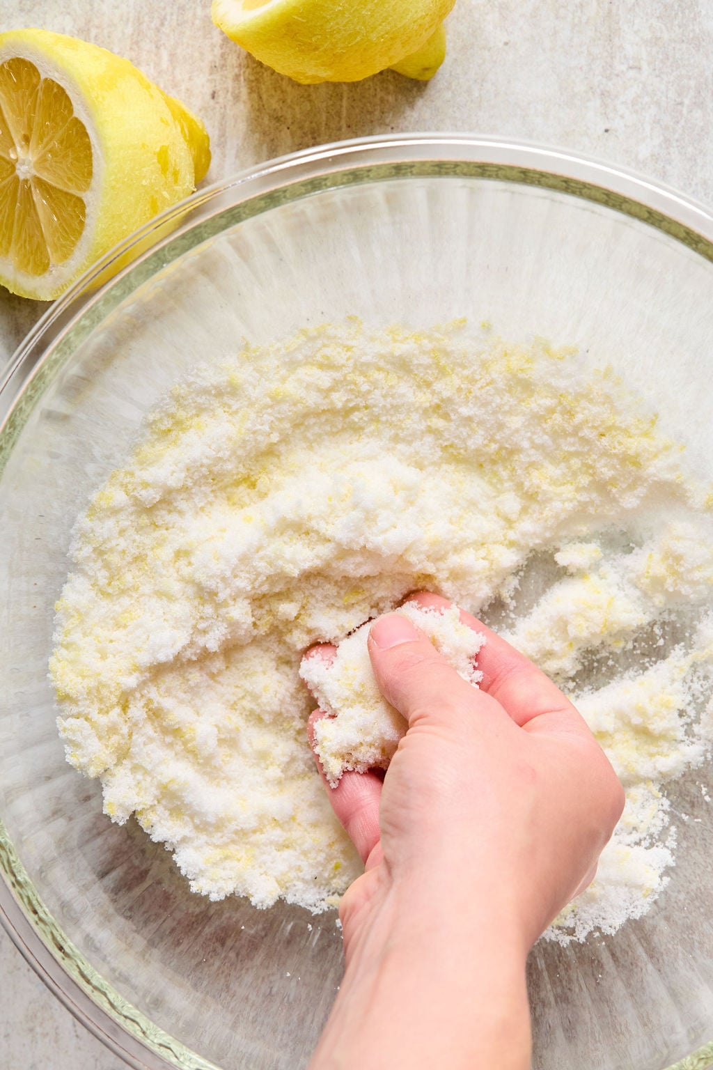 A hand mixing sugar and lemon zest in a glass bowl with halved lemons nearby on a light surface.