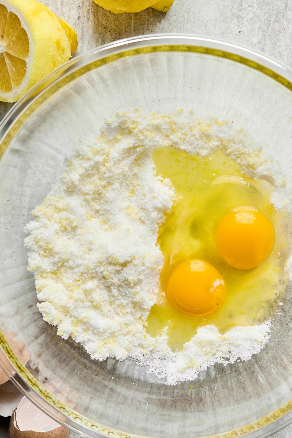 A glass bowl containing two cracked eggs and flour mixture, with lemon and eggshells visible nearby on a light surface.