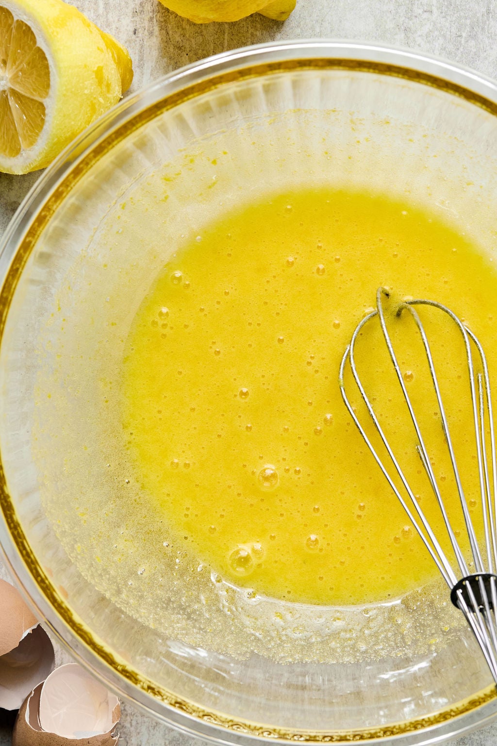 A clear glass bowl with a yellow mixture being whisked, surrounded by broken eggshells and a partially cut lemon.