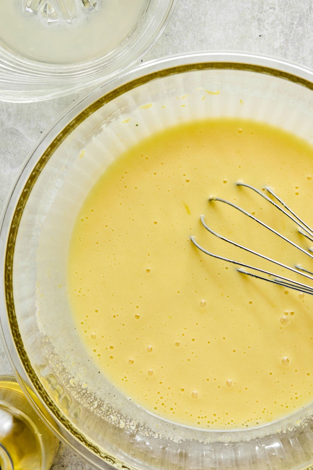 A glass bowl of yellow batter with a metal whisk partially submerged, placed on a light surface.