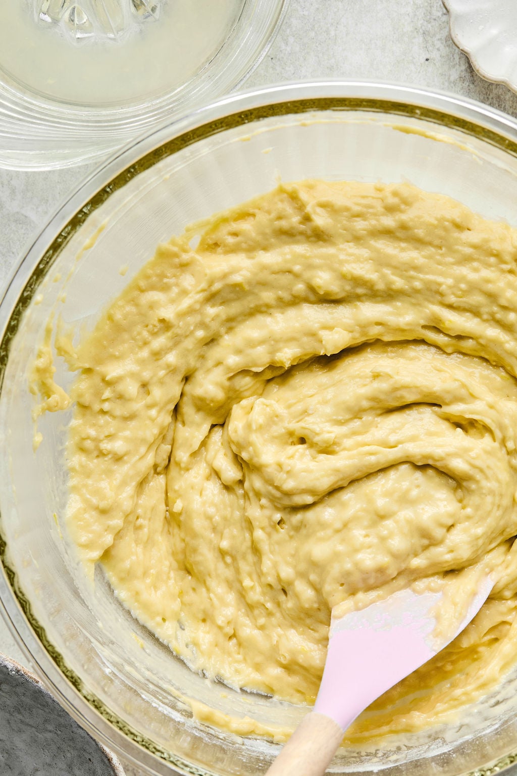 A glass bowl filled with thick, pale yellow batter being mixed with a pink spatula.