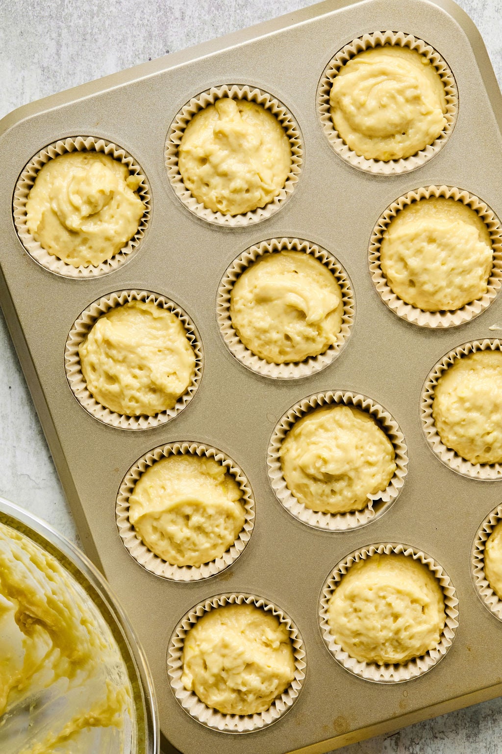 A muffin tin lined with paper cups is filled with raw yellow batter, ready to be baked. A mixing bowl with leftover batter is visible in the bottom left corner.