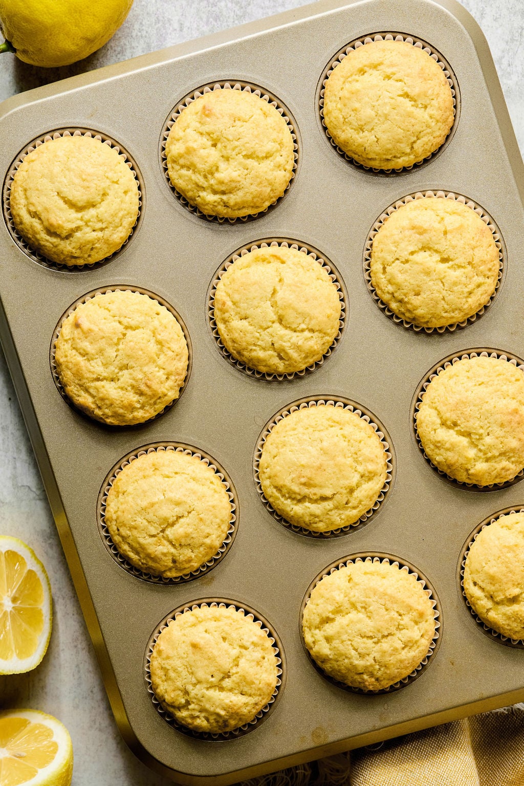 A muffin tin filled with twelve baked lemon muffins, with sliced lemons beside the tin on a light surface.
