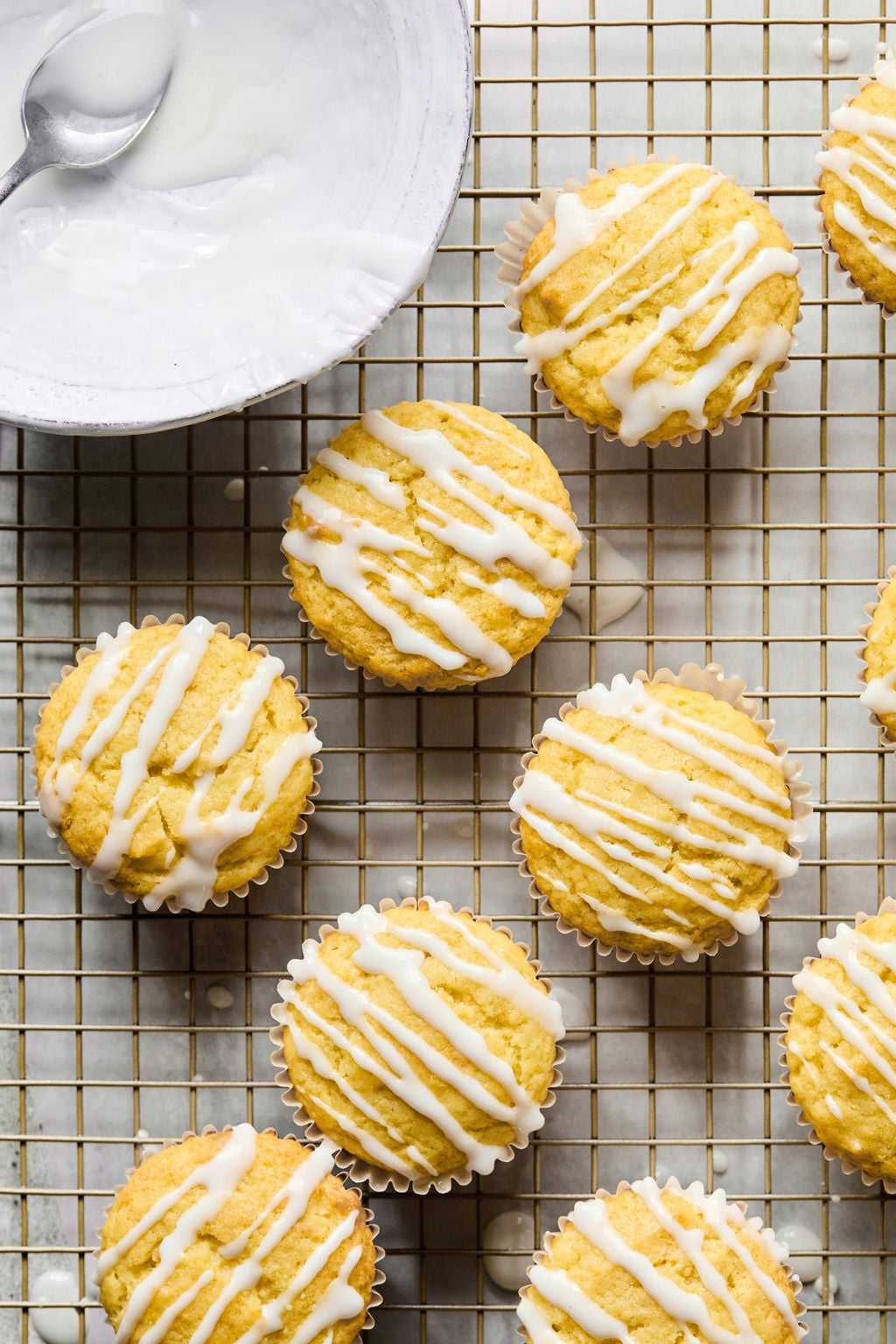 Lemon muffins with white icing drizzle arranged on a cooling rack, with a bowl and spoon of icing nearby.