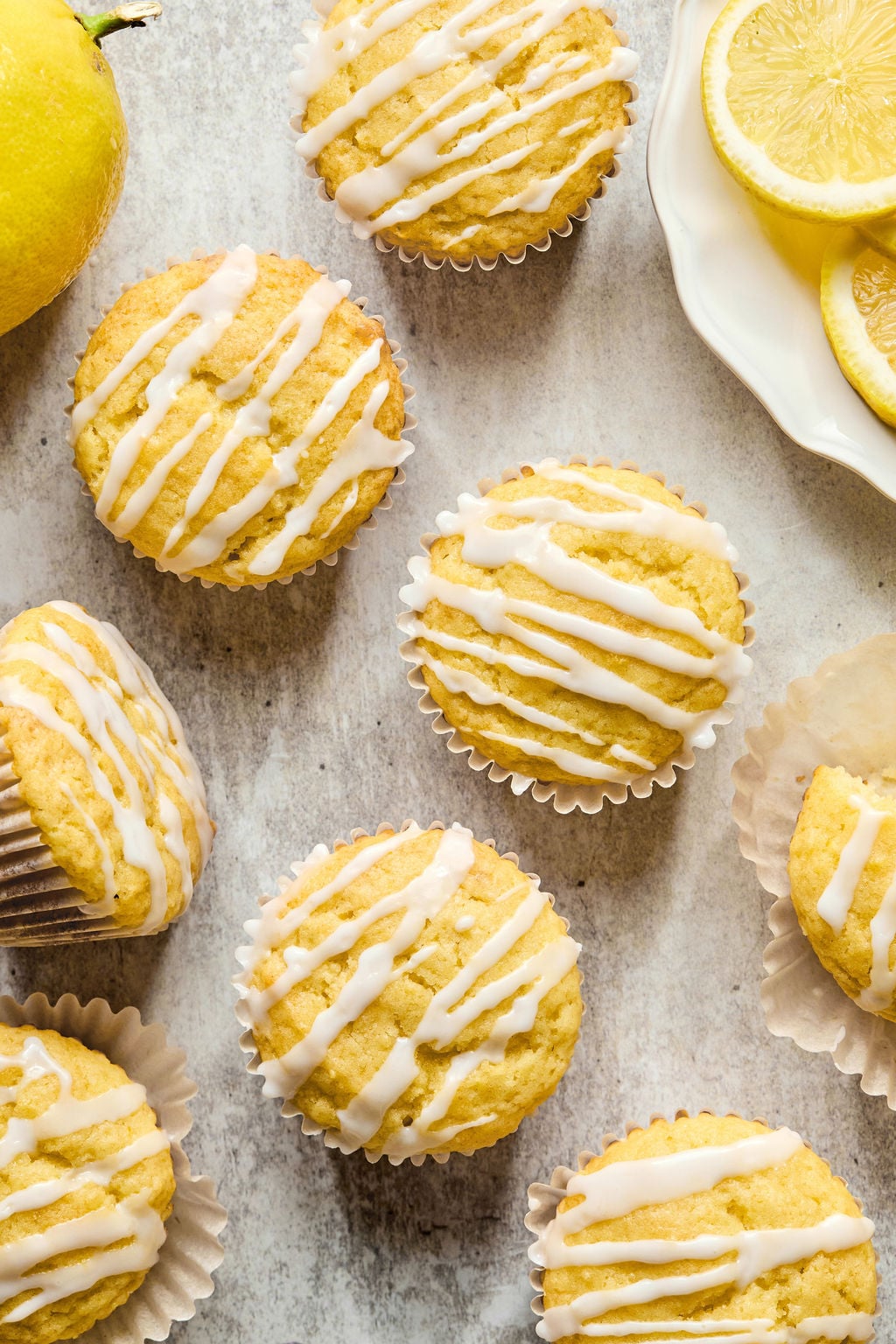 Lemon muffins with white icing drizzle arranged on a light surface, next to a sliced lemon and a whole lemon.