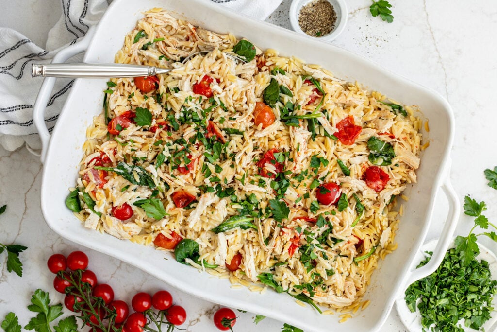 A white baking dish filled with orzo pasta, shredded chicken, spinach, cherry tomatoes, and herbs, with a serving spoon. Fresh parsley and tomatoes are arranged nearby.