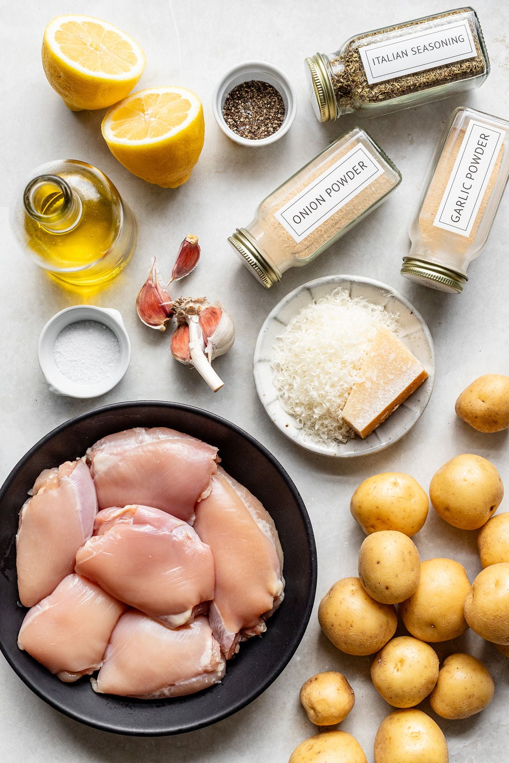 Raw chicken thighs, baby potatoes, lemon halves, garlic, olive oil, grated Parmesan, salt, pepper, Italian seasoning, onion powder, and garlic powder arranged on a countertop.