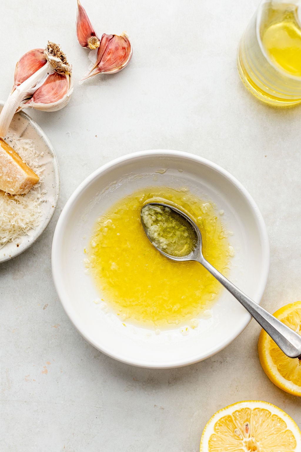 A white bowl with a spoon contains a mixture of oil, grated garlic, and lemon juice. Surrounding the bowl are garlic cloves, Parmesan cheese, lemon slices, and a jar of oil.