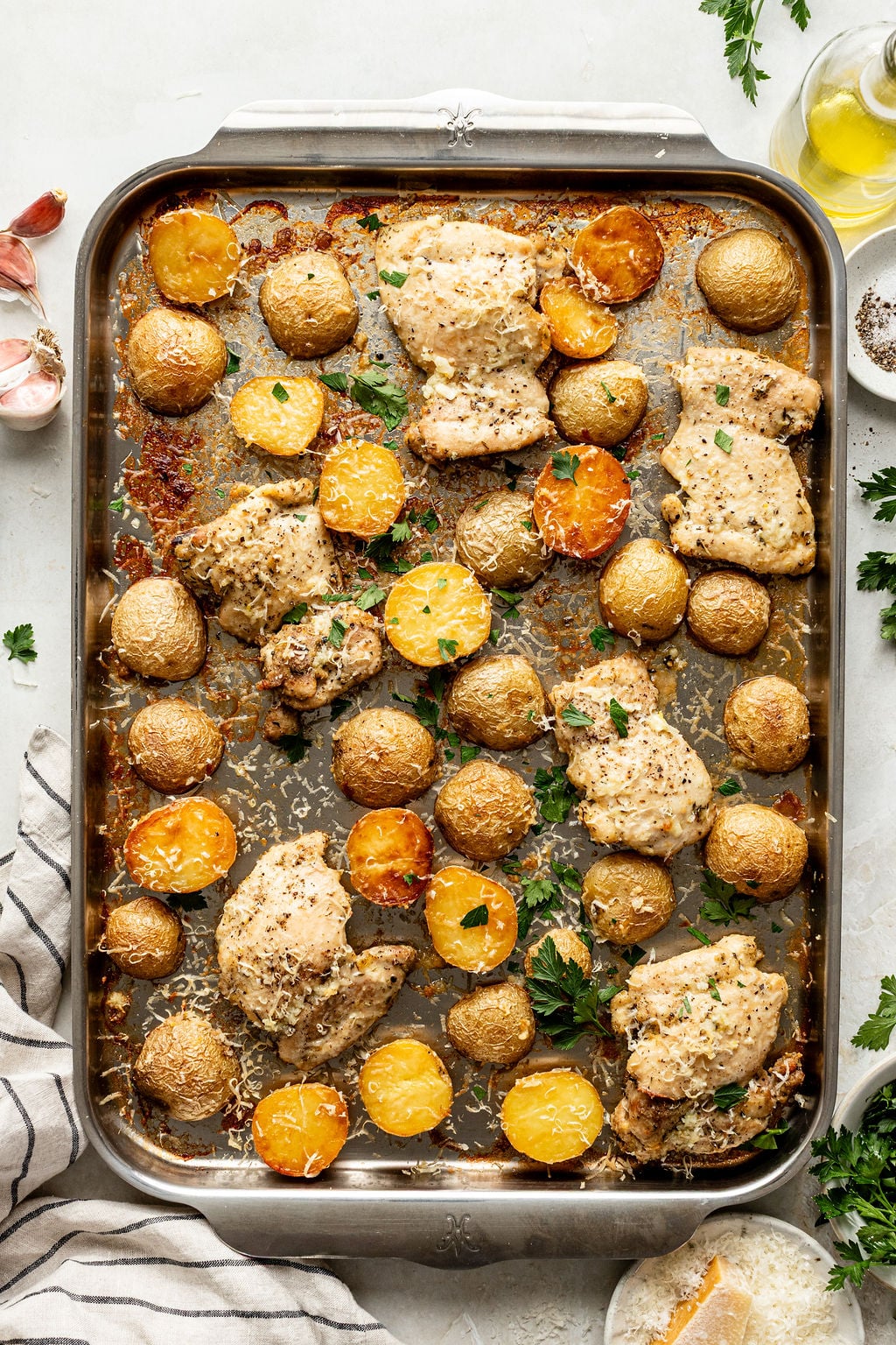 Baked chicken thighs and baby potatoes on a metal sheet pan, garnished with herbs and grated cheese.