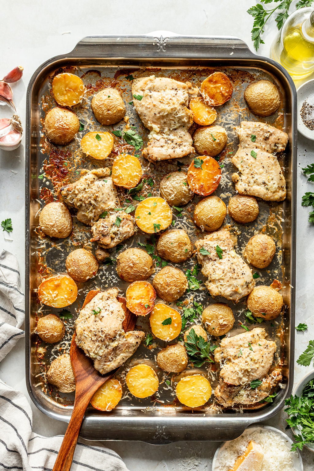 Baked chicken thighs and baby potatoes arranged on a metal baking tray, garnished with herbs. A wooden spoon rests in the corner.