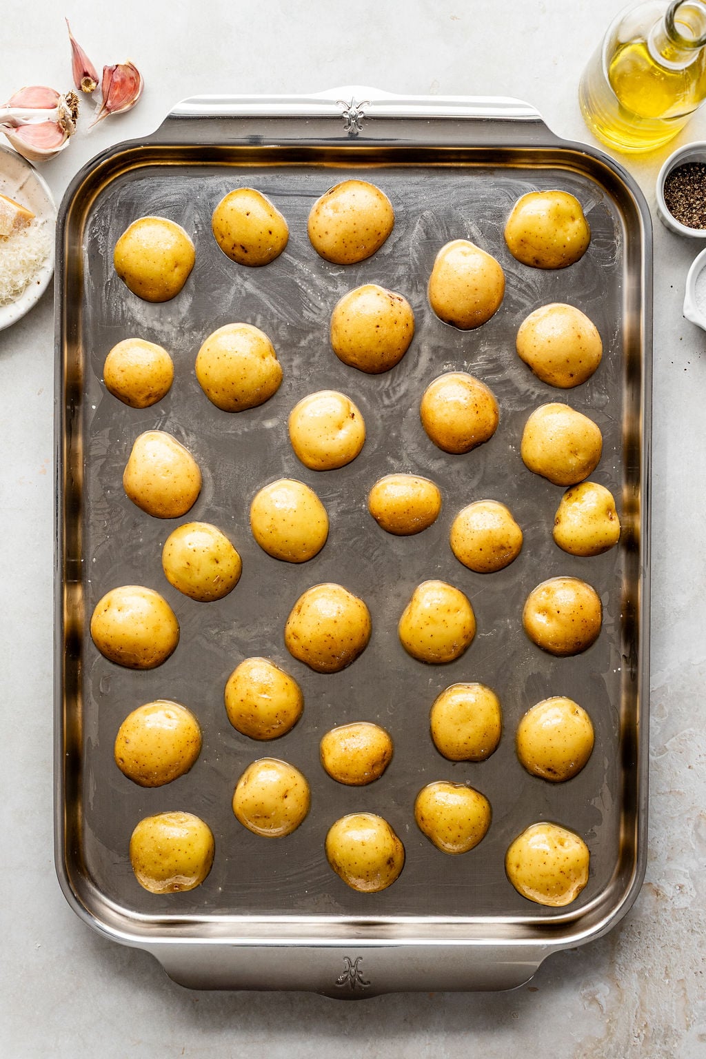 Raw baby potatoes arranged on a metal baking sheet, ready to be roasted. Garlic, olive oil, salt, and pepper are visible nearby.