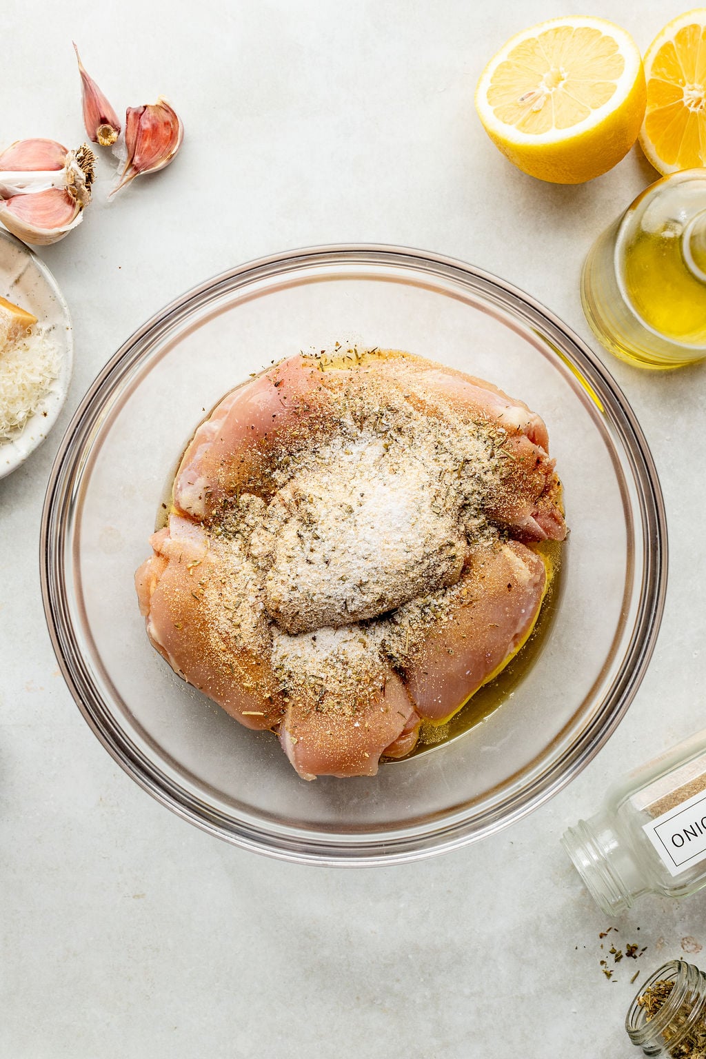 Raw chicken breasts in a glass bowl topped with spices, and oil, surrounded by garlic, lemon halves, olive oil, and seasonings on a countertop.