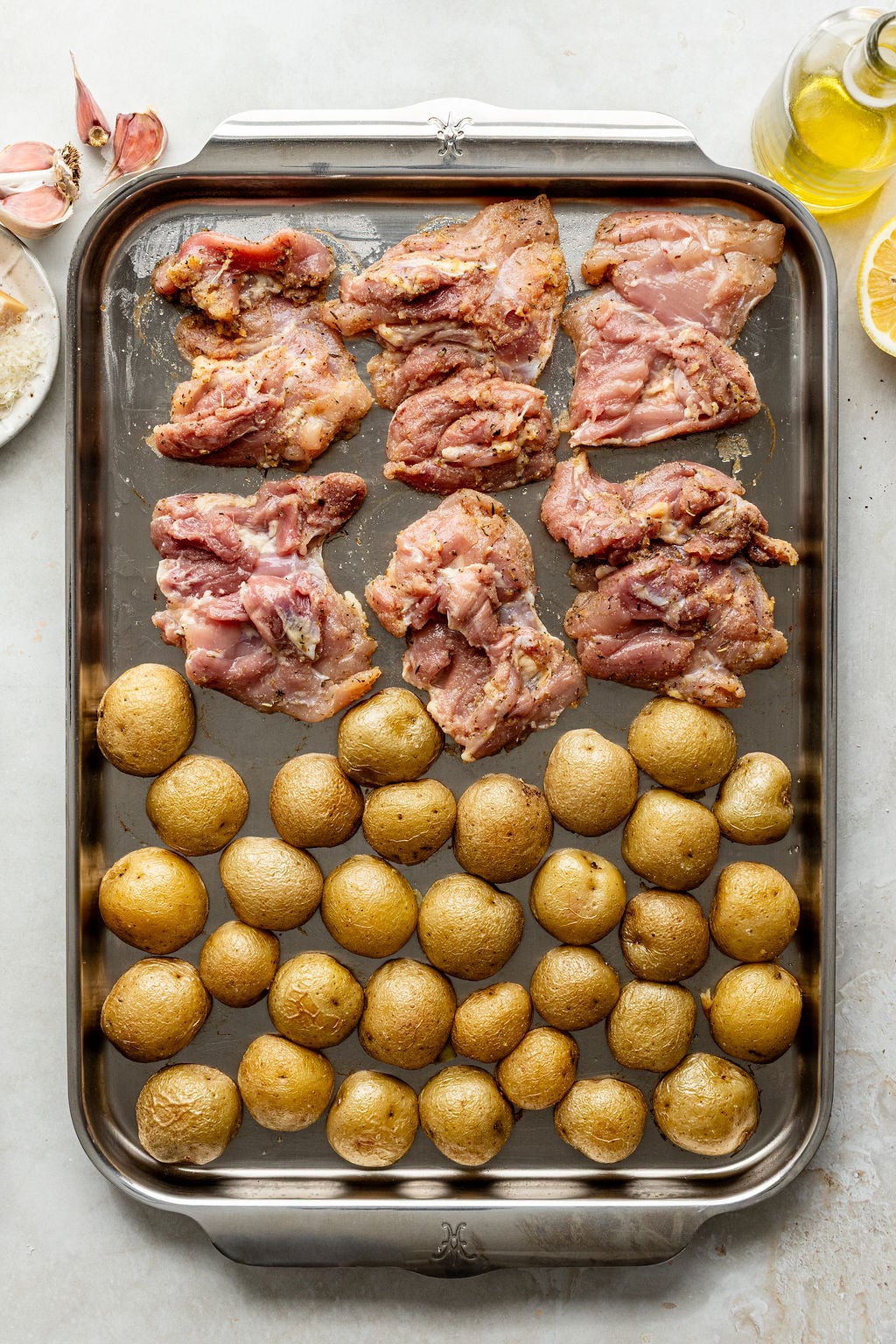 A metal baking sheet with raw seasoned chicken thighs and baby potatoes arranged in rows, ready to be cooked.
