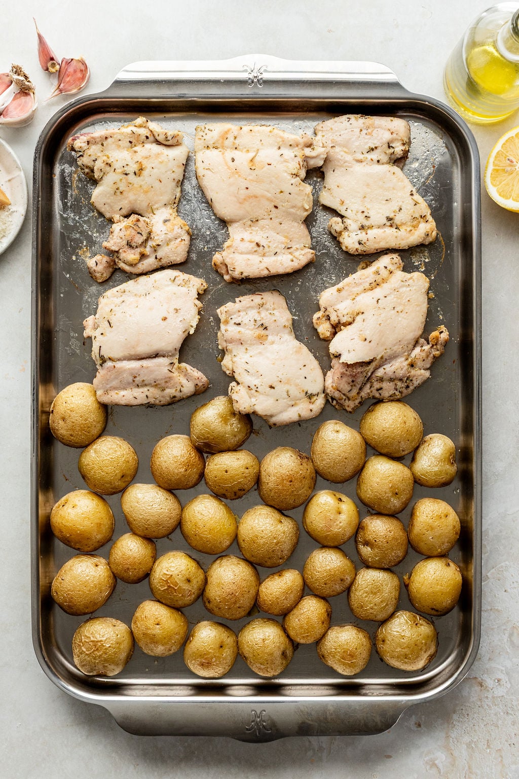 A baking tray with seasoned raw chicken thighs in the top half and baby potatoes in the bottom half, ready to be cooked.